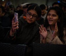 Two girls are taking a selfie with their cell phones while sitting in a crowd.
