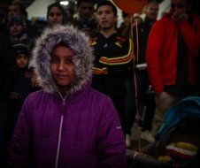 A girl in a purple jacket with a fur hood is standing in front of a crowd of people.