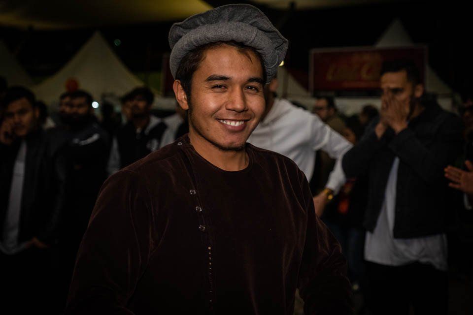 A man wearing a hat and a brown jacket is smiling in front of a crowd of people.