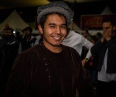 A man wearing a hat and a brown jacket is smiling in front of a crowd.