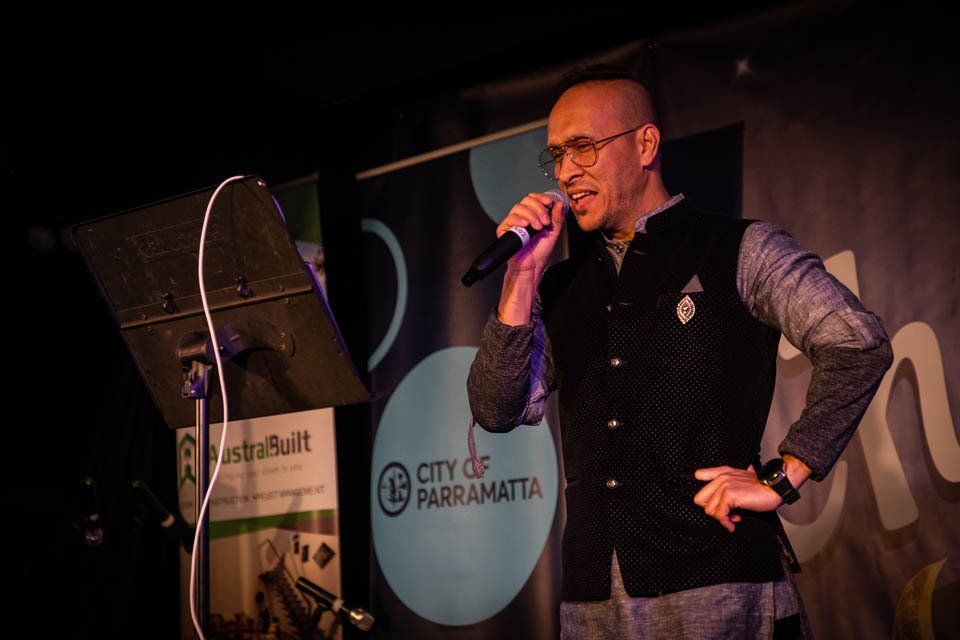 A man is singing into a microphone in front of a sign that says city of parramatta