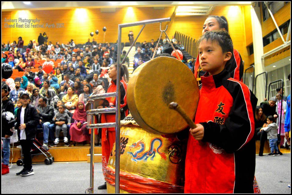 A boy in a red jacket is playing a drum in front of a crowd