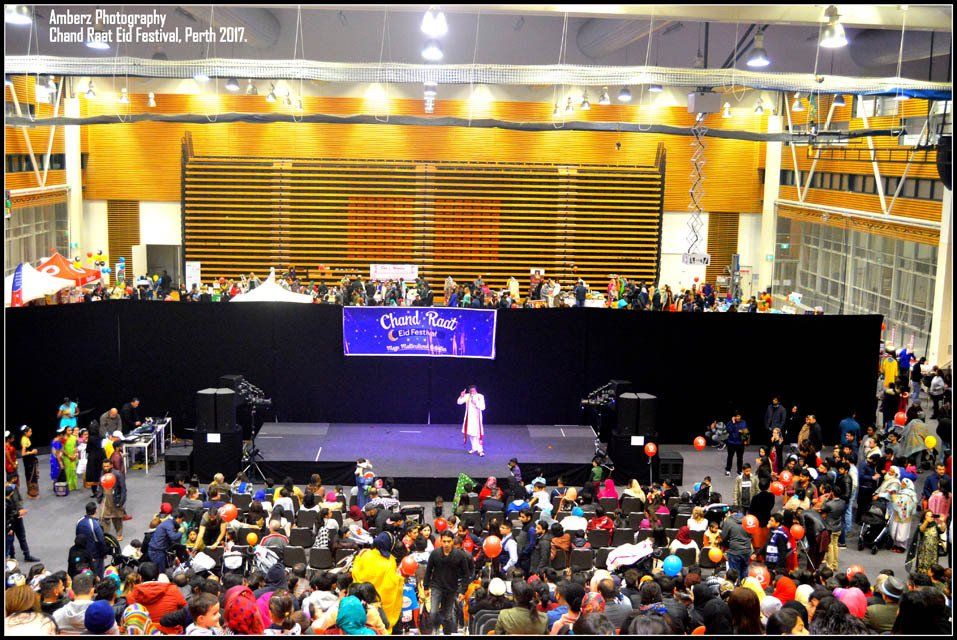 A crowd of people are gathered in a large room with a stage and a sign that says coolest kid in the world