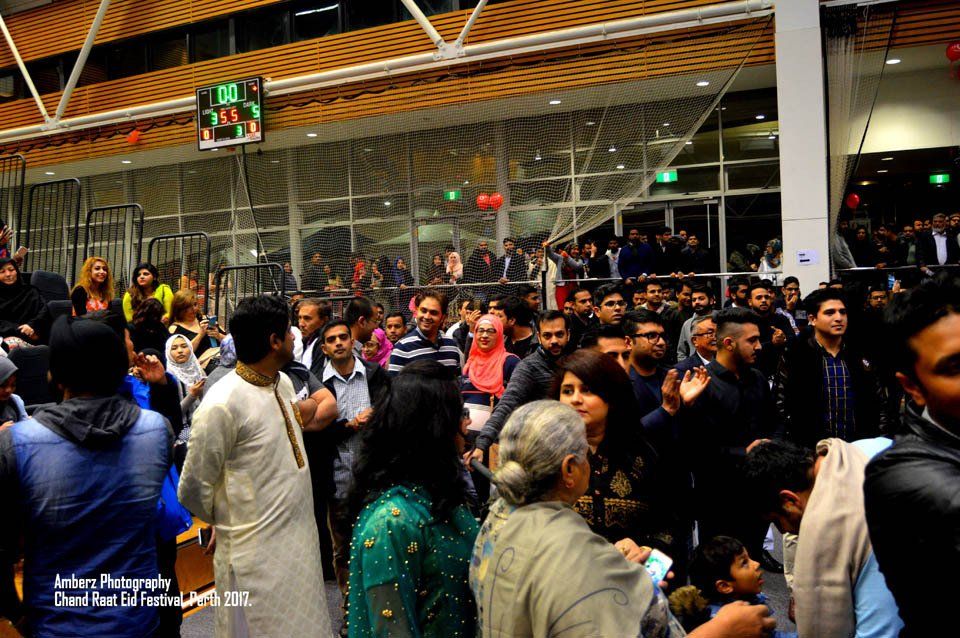 A large group of people are standing in a room with a scoreboard in the background.