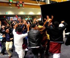 A group of people are dancing in a room with a scoreboard in the background.