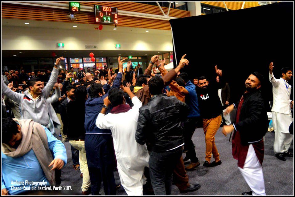 A group of men are dancing in a room with a scoreboard in the background