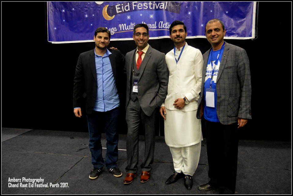 Four men are posing for a picture in front of a sign that says eid festival