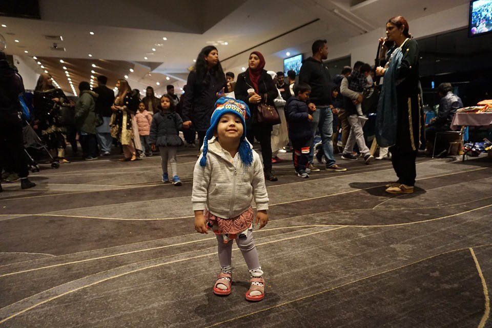 A little girl wearing a blue hat is standing in a crowded room.