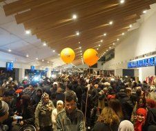 A crowd of people are standing in a room with balloons hanging from the ceiling.