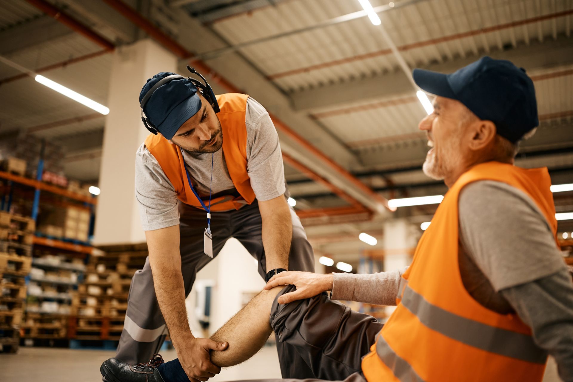 Man helping another man who has injured his leg in a warehouse. Both are wearing orange vests and work clothes.