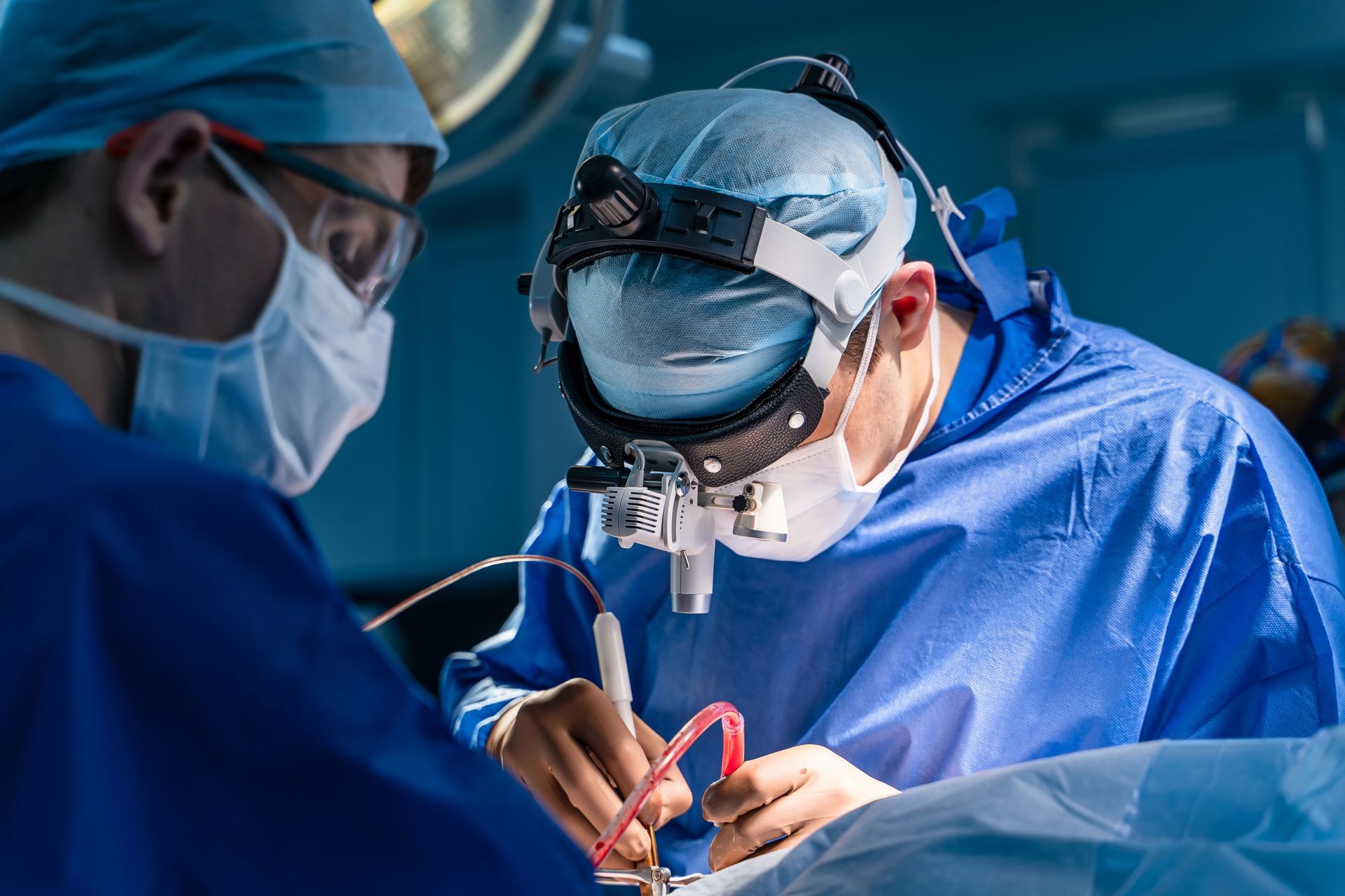 Surgeons in blue scrubs operating in a surgical suite. One uses a surgical microscope.