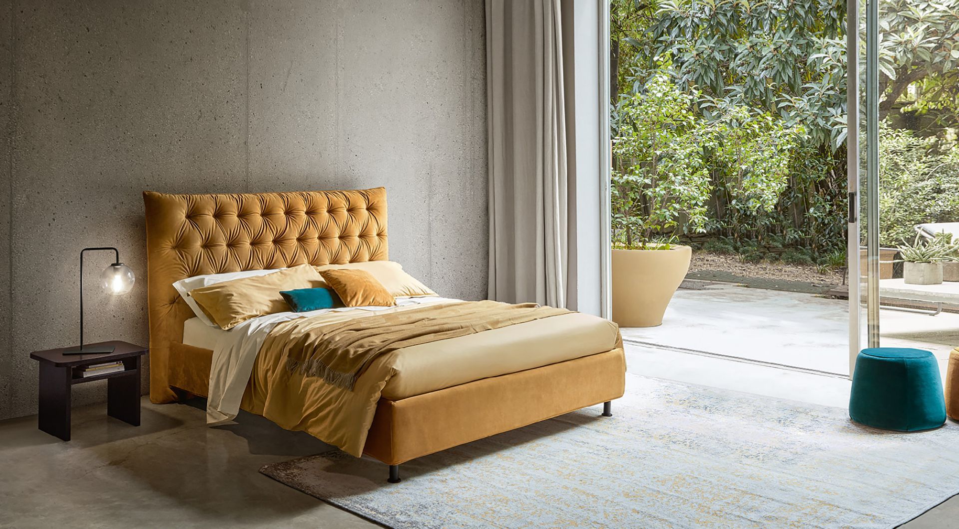 Letto imbottito color senape con cuscini, accanto a un comodino nero con lampada e una grande finestra.