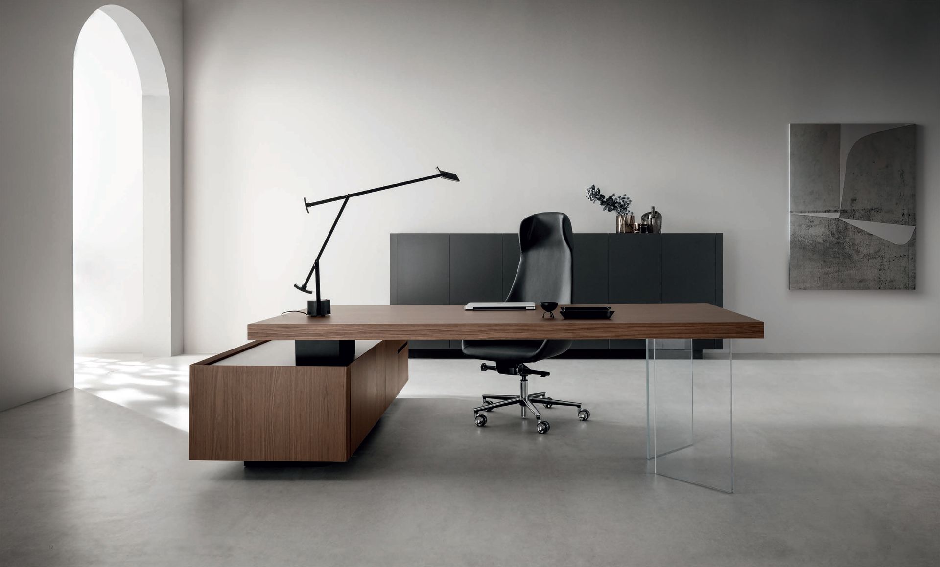 Ufficio moderno con scrivania in legno, sedia nera, lampada e mobiletto portaoggetti contro una parete bianca.