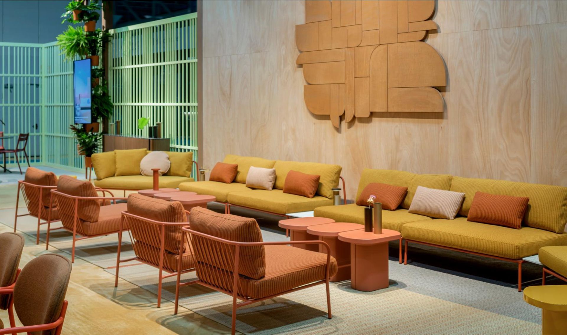 Accogliente salotto con divani color senape, sedie intrecciate, tavolini in legno e luci calde su una parete con finitura materica.