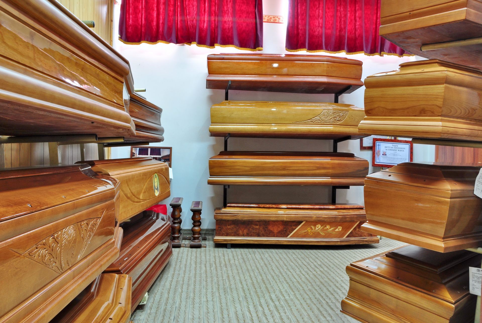 Coffin display in a showroom, wooden caskets on shelves. Red curtains in background.