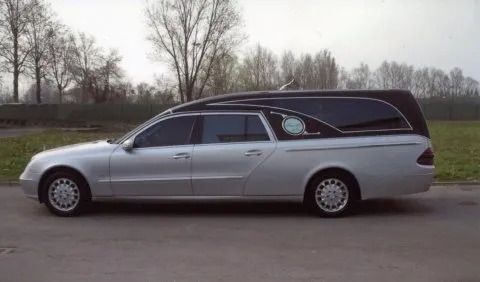 Silver hearse parked on a road, with a black extended roof and tinted windows.