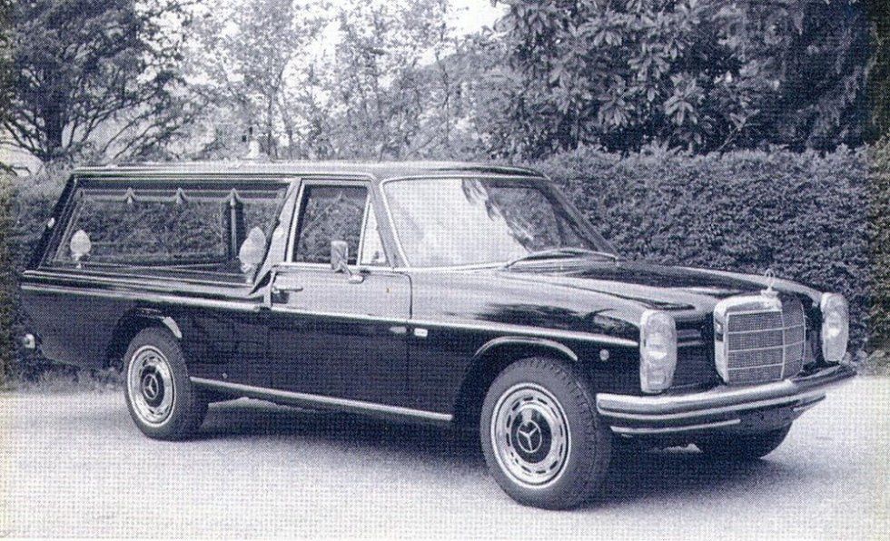 Black Mercedes hearse parked on a paved surface, with an enclosed rear compartment. Trees in the background.