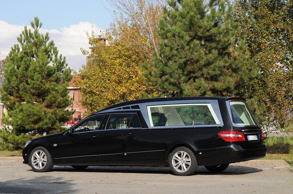Black hearse parked outdoors, near trees and a building on a partly sunny day.