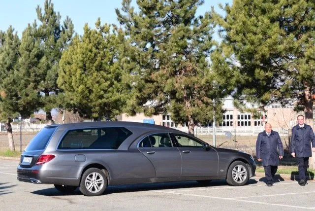 Dark gray hearse parked outdoors, two men in coats standing beside it. Trees and building in background.