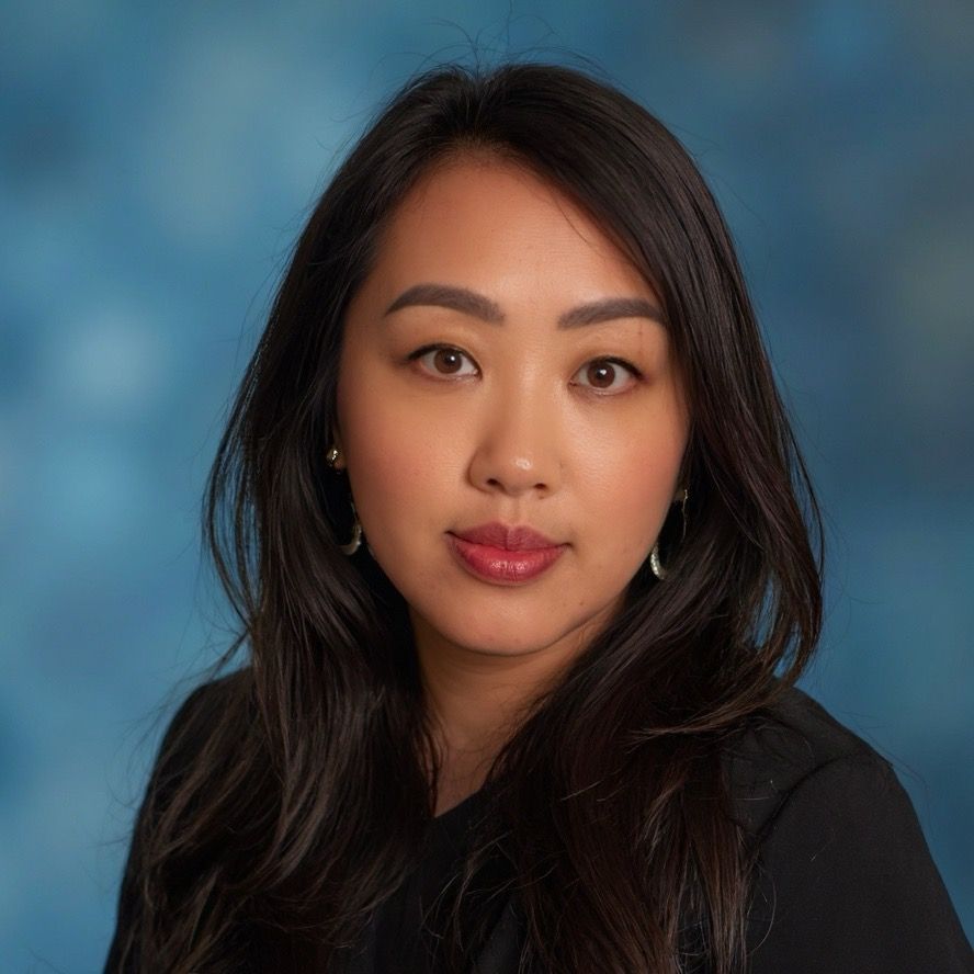 Woman with long dark hair, wearing a black shirt, looking at the camera. Blue background.