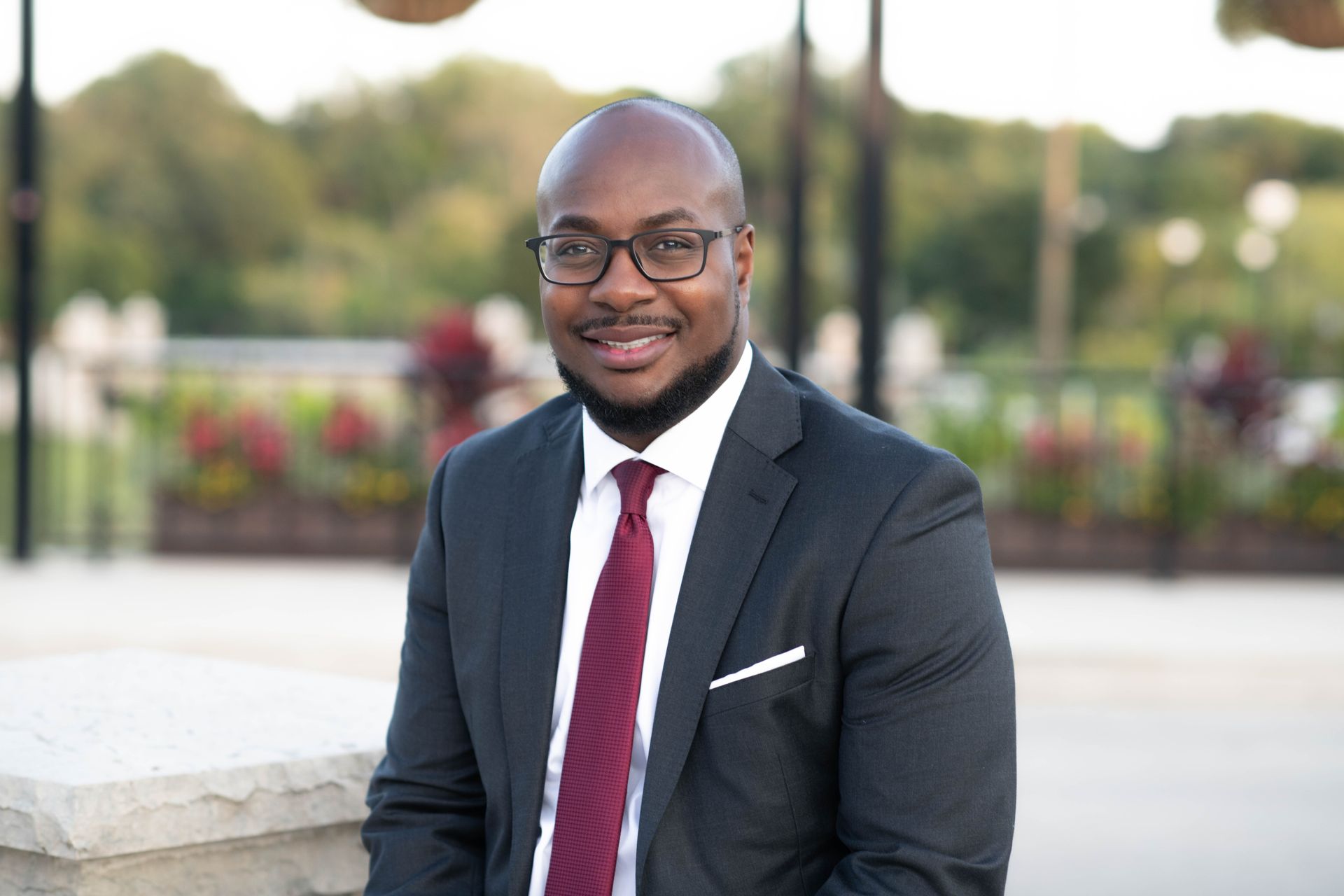 A professional portrait of a smiling person wearing a dark suit, white shirt, and a burgundy tie outdoors.