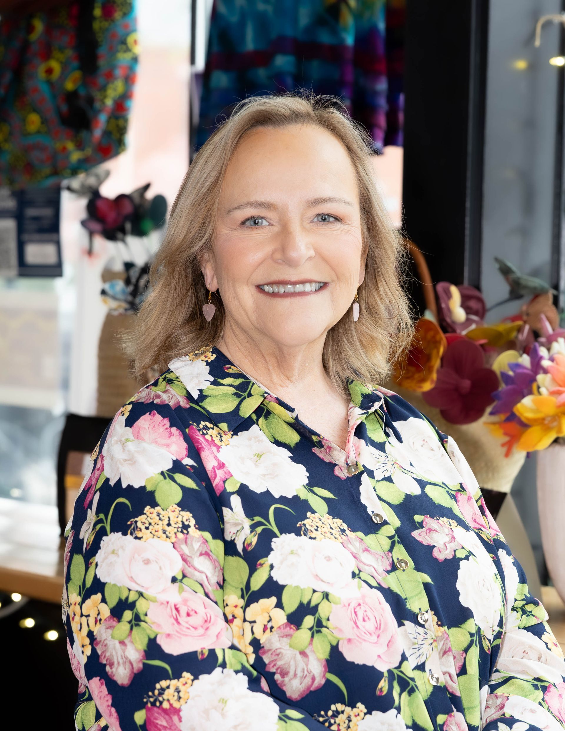 A smiling person wearing a floral print button-down shirt stands in a brightly lit indoor space with blurred retail items.