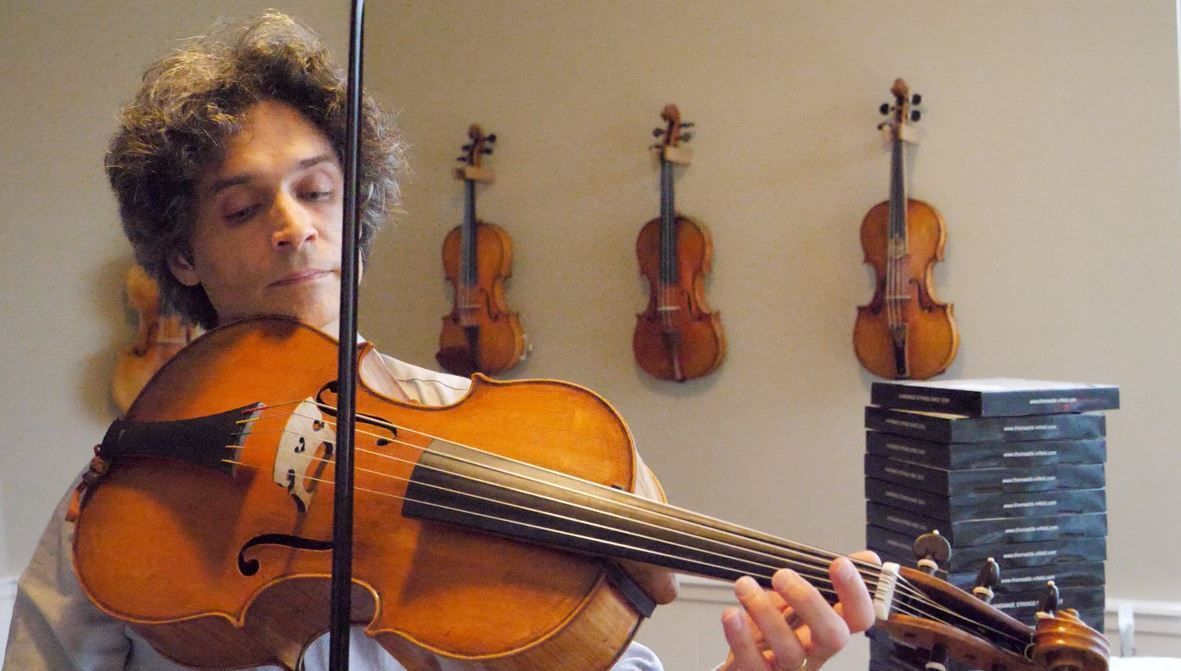 A man is playing a violincello da spalla in front of a wall of violins