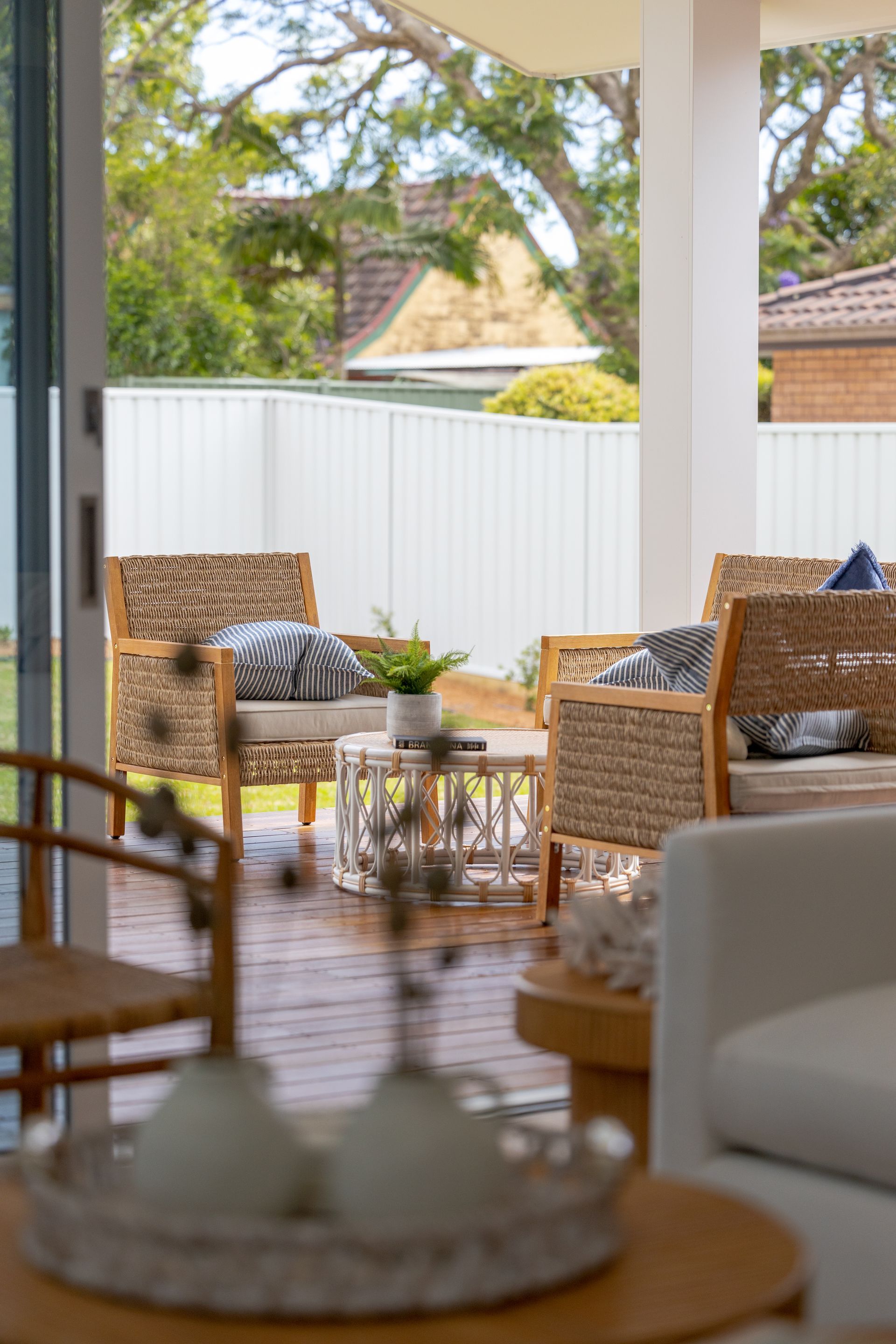 Outdoor living space: wicker chairs, coffee table, deck. White fence and greenery in the background. — Blue Horizon Projects in Bateau Bay, NSW
