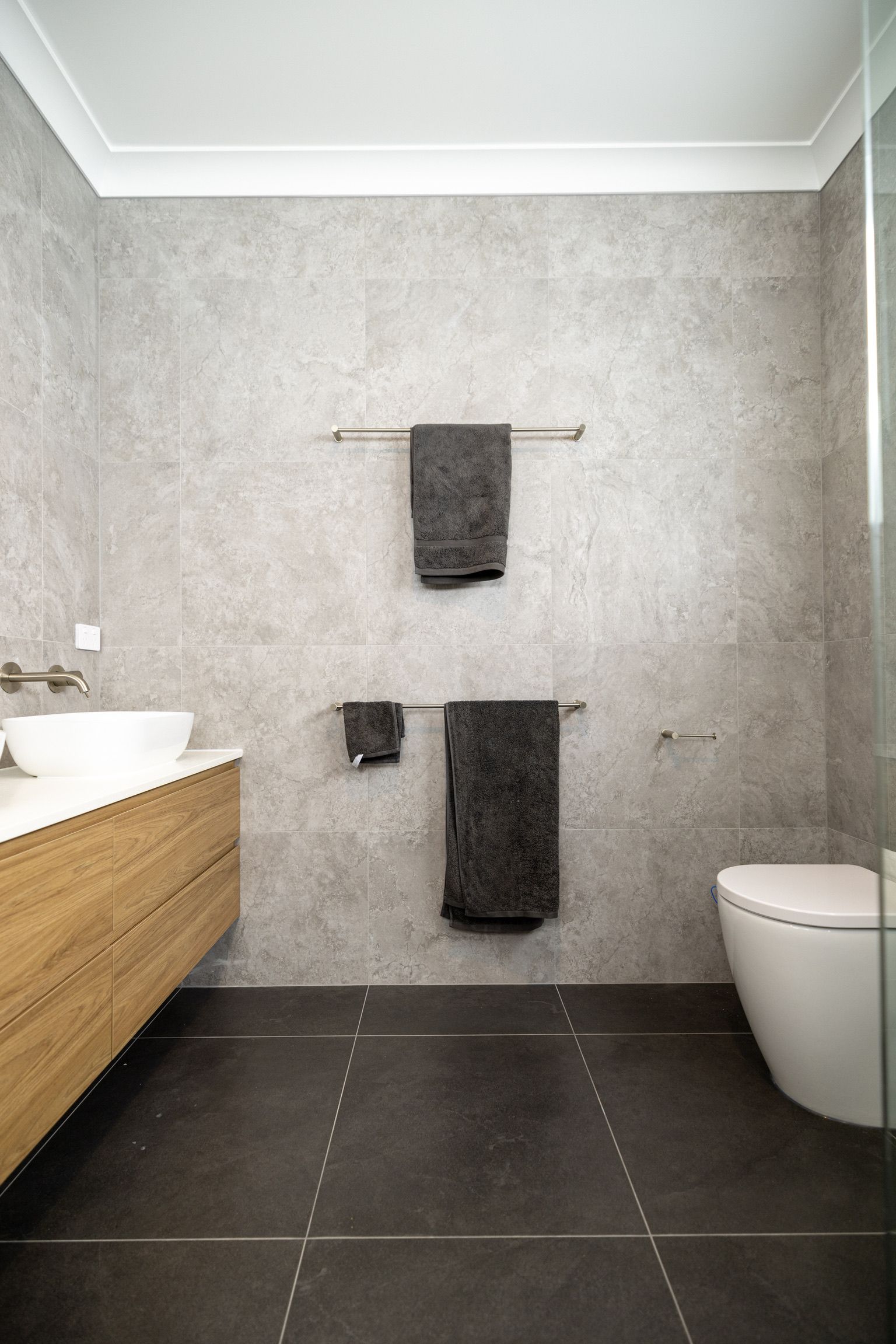 Bathroom with gray tiled walls, wooden vanity, two towel racks with dark towels, and a white toilet. — Blue Horizon Projects in Bateau Bay, NSW