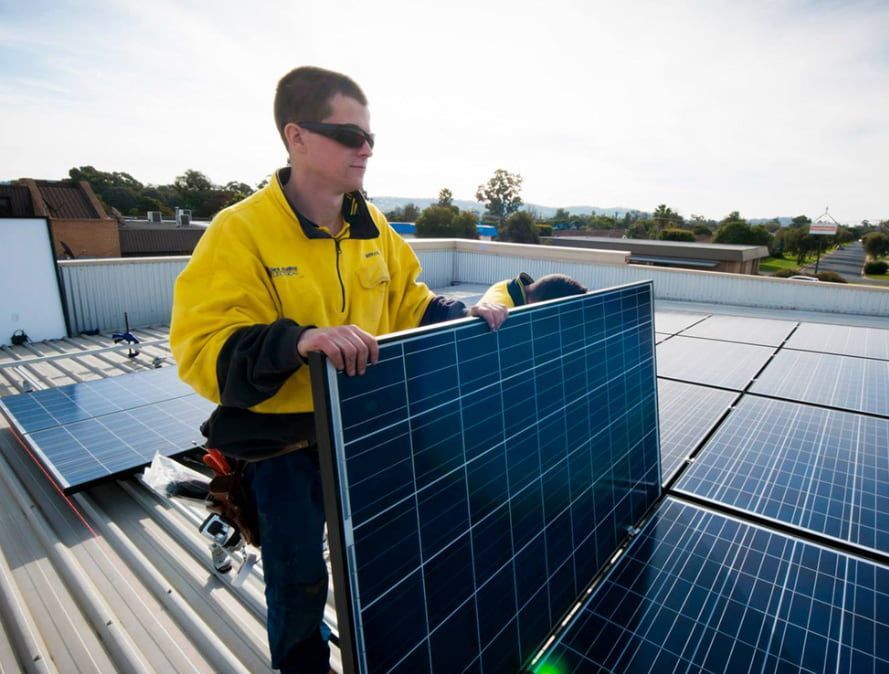 A Man in a Yellow Jacket is Holding a Solar Panel on Top of a Roof — Des Mullins Electrical & Solar In Wagga Wagga, NSW