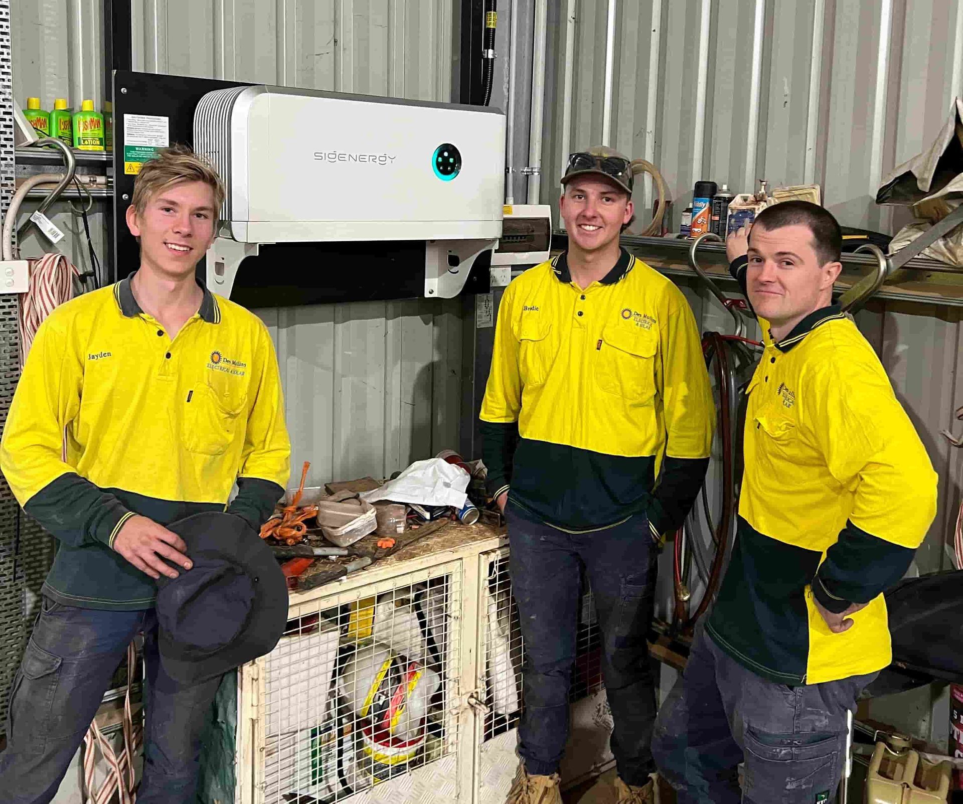 Three men in yellow shirts are standing next to each other in a garage — Des Mullins Electrical & Solar In Wagga Wagga, NSW