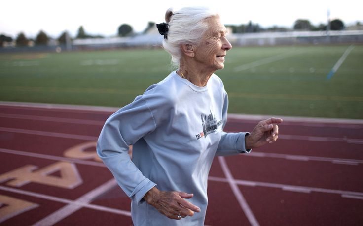 Elderly woman running on a track, smiling. Wearing blue shirt, white hair. Stadium in the background.