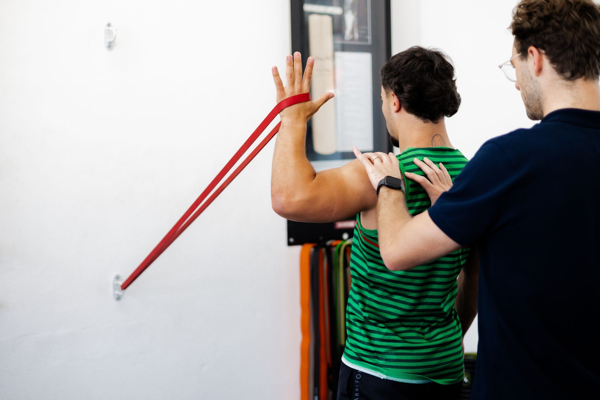 Man doing shoulder exercise with red resistance band, assisted by another man, near a white wall.