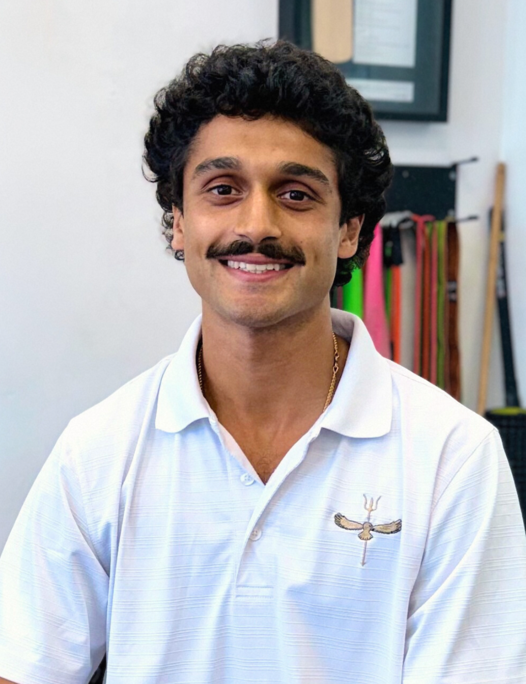Man with curly hair and mustache smiles, wearing a white polo shirt with a small emblem.