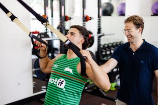 Man using TRX suspension straps, guided by an instructor in a gym setting.