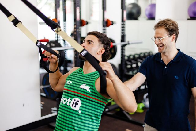 Man using TRX suspension straps, guided by an instructor in a gym setting.