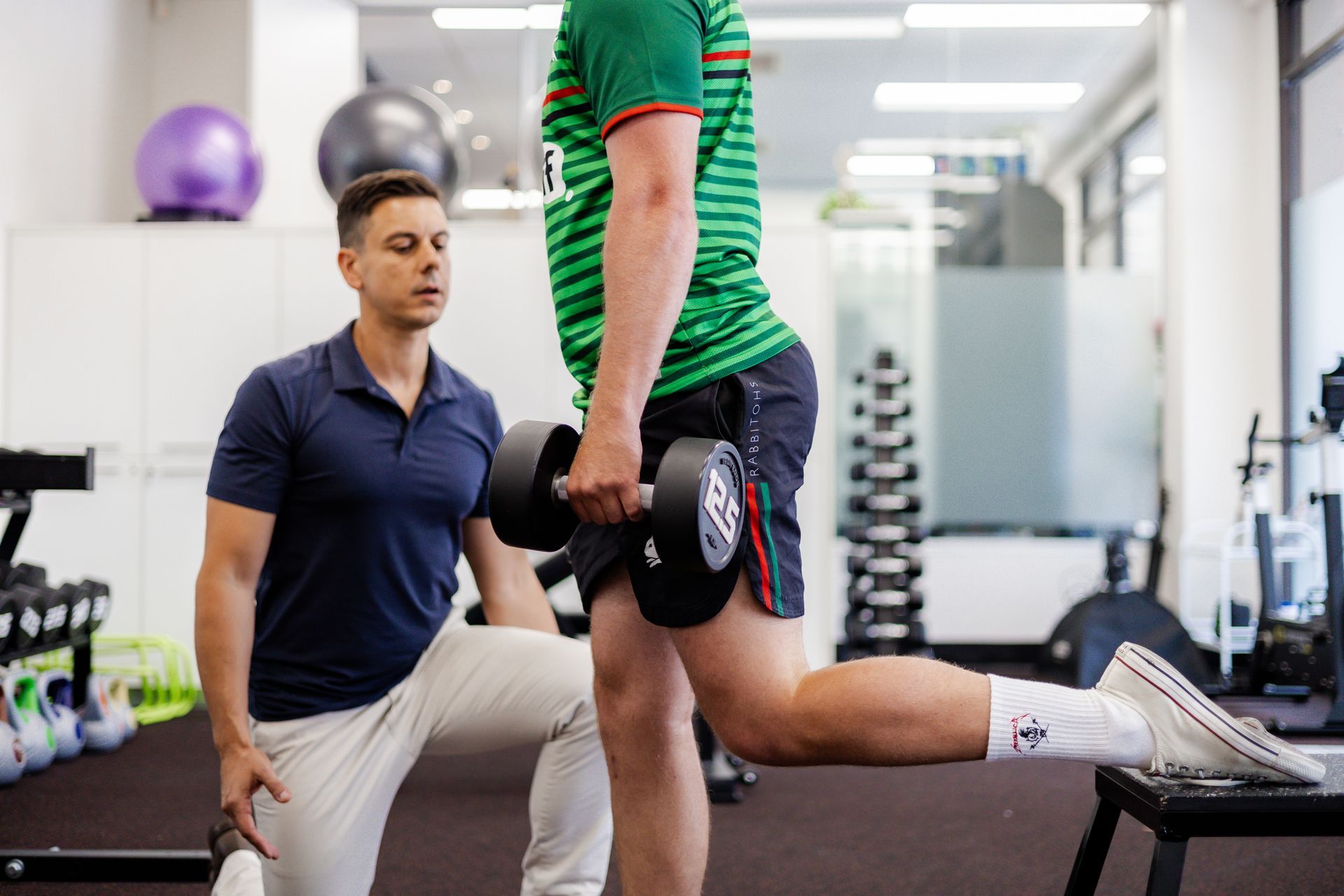 Man performing a dumbbell Bulgarian split squat under the supervision of a physical therapist in a gym.
