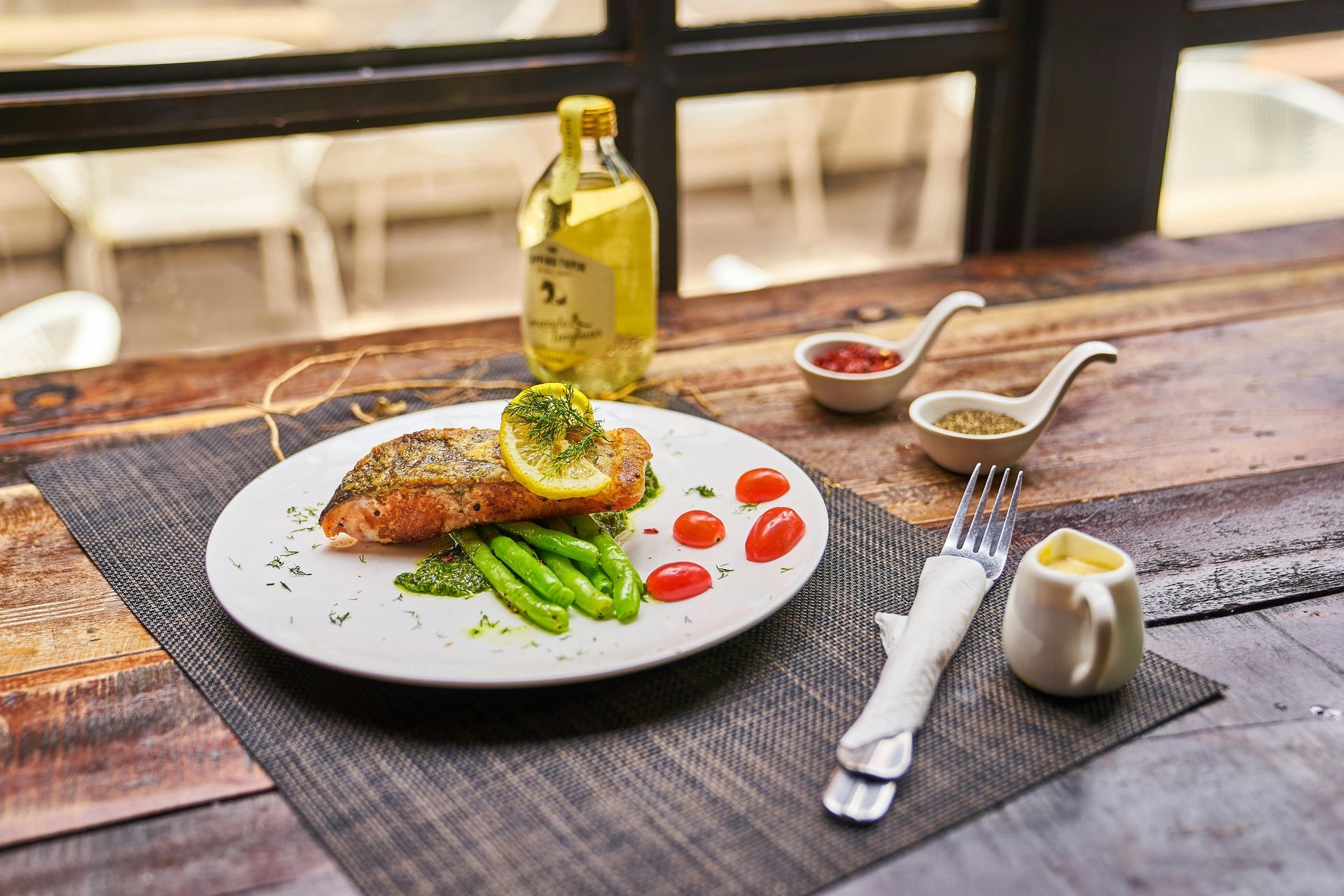 A plated salmon fillet with green beans and cherry tomatoes, served on a table with side sauces and a glass oil bottle.