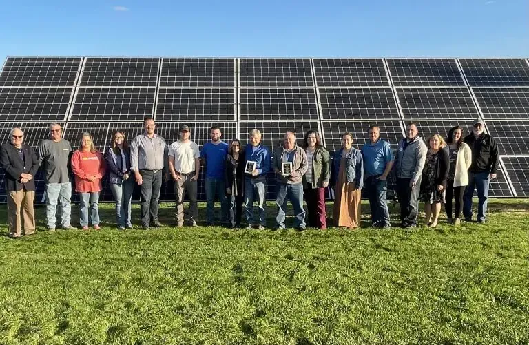 A diverse group of people standing in a line in front of a large solar panel array on a grassy field.