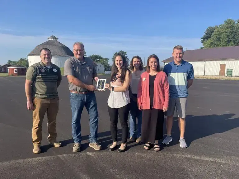 Six people stand in a line on a paved lot outdoors, with a man handing a small award to a woman in the center.