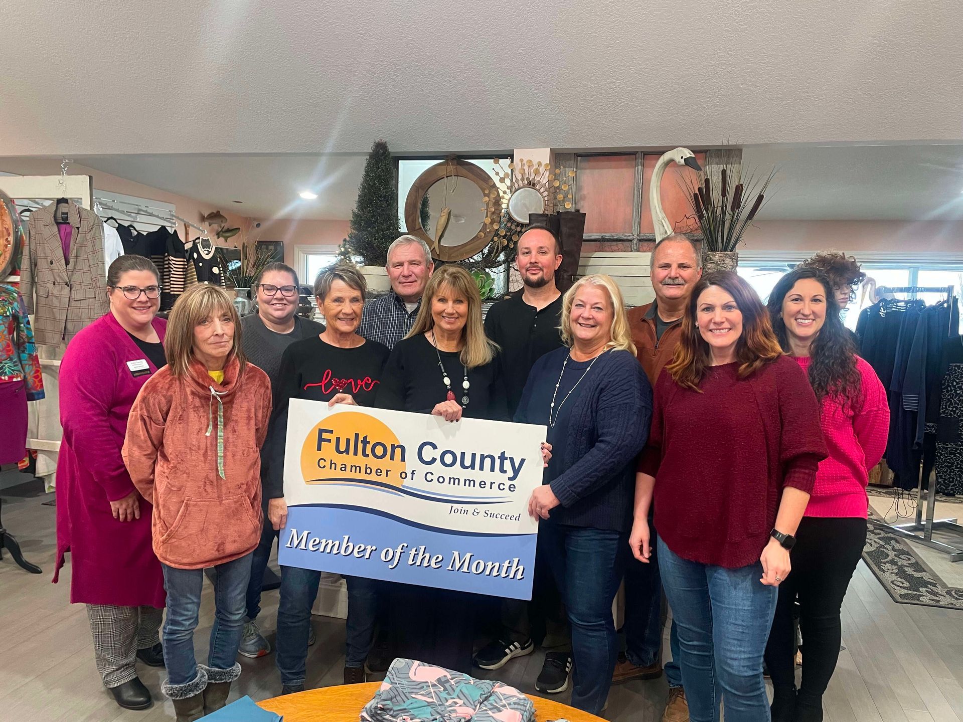 A group of people standing indoors, holding a "Fulton County Chamber of Commerce Member of the Month" sign.