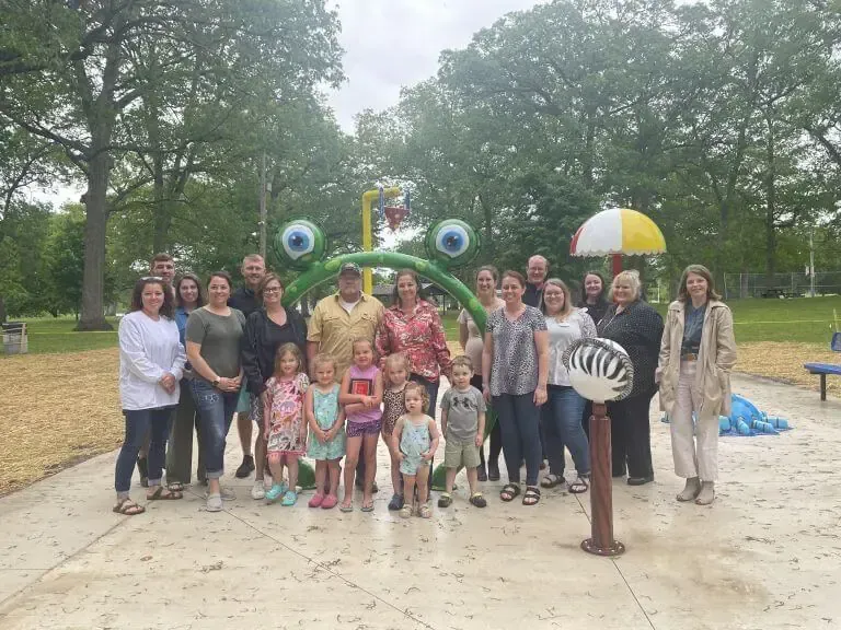 A group of adults and children stand in front of a green frog-themed water feature at a park with trees in the background.