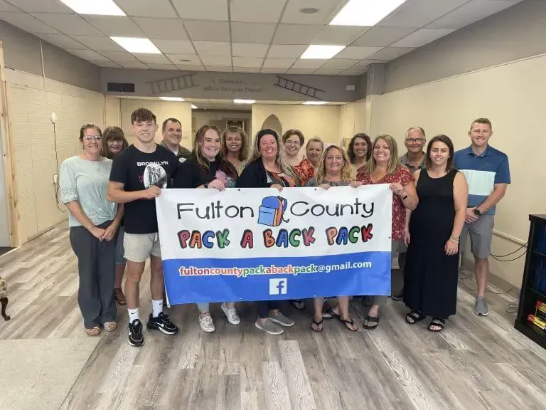 A group of people holding a banner for Fulton County Pack a Back Pack in a community room with wood-look flooring.