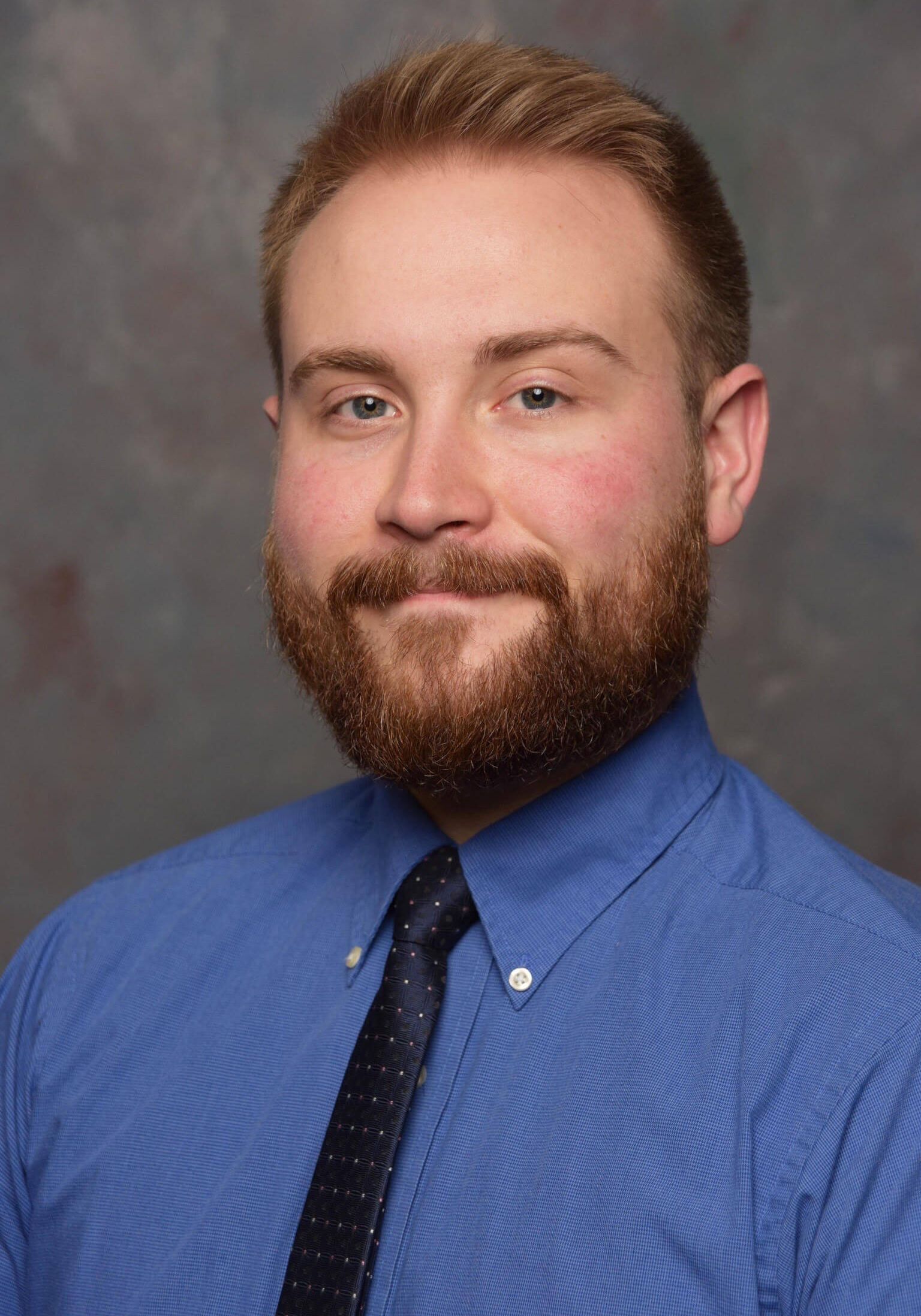 Professional headshot of a person with a beard, wearing a blue button-down shirt and a dark tie against a gray background.