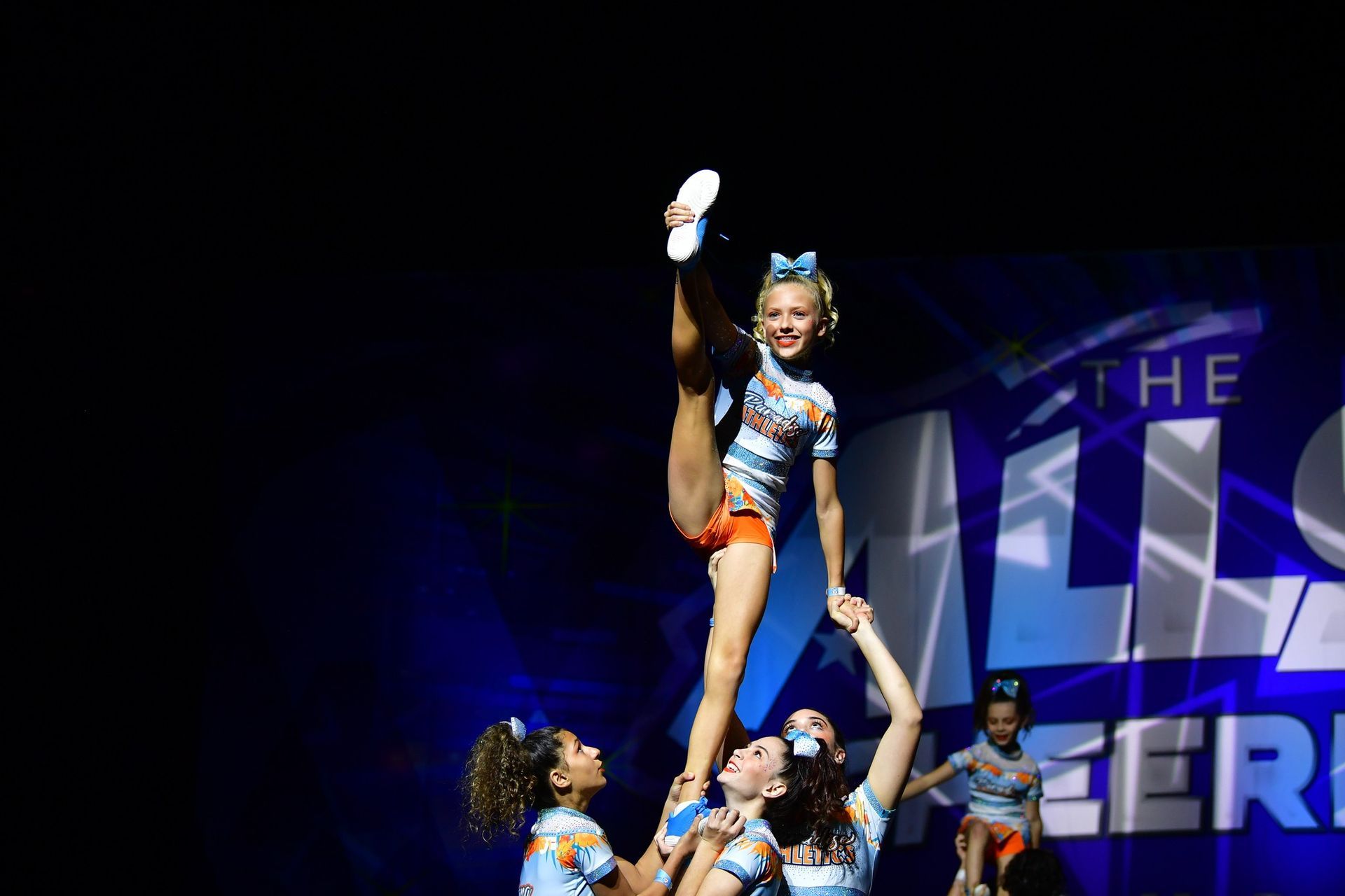 A cheerleader in a light blue and orange uniform stands in a heel stretch on the hands of teammates on a stage.