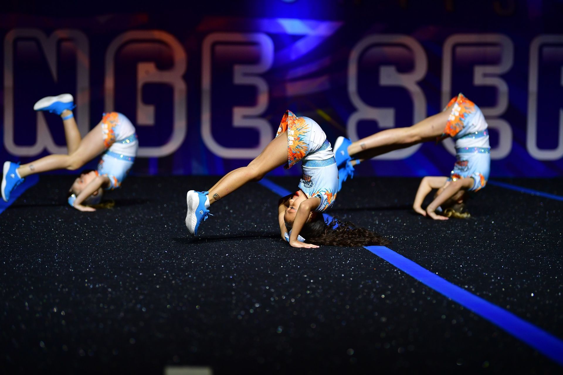 Three cheerleaders in light blue uniforms perform synchronized handstands on a dark, speckled mat under stage lights.