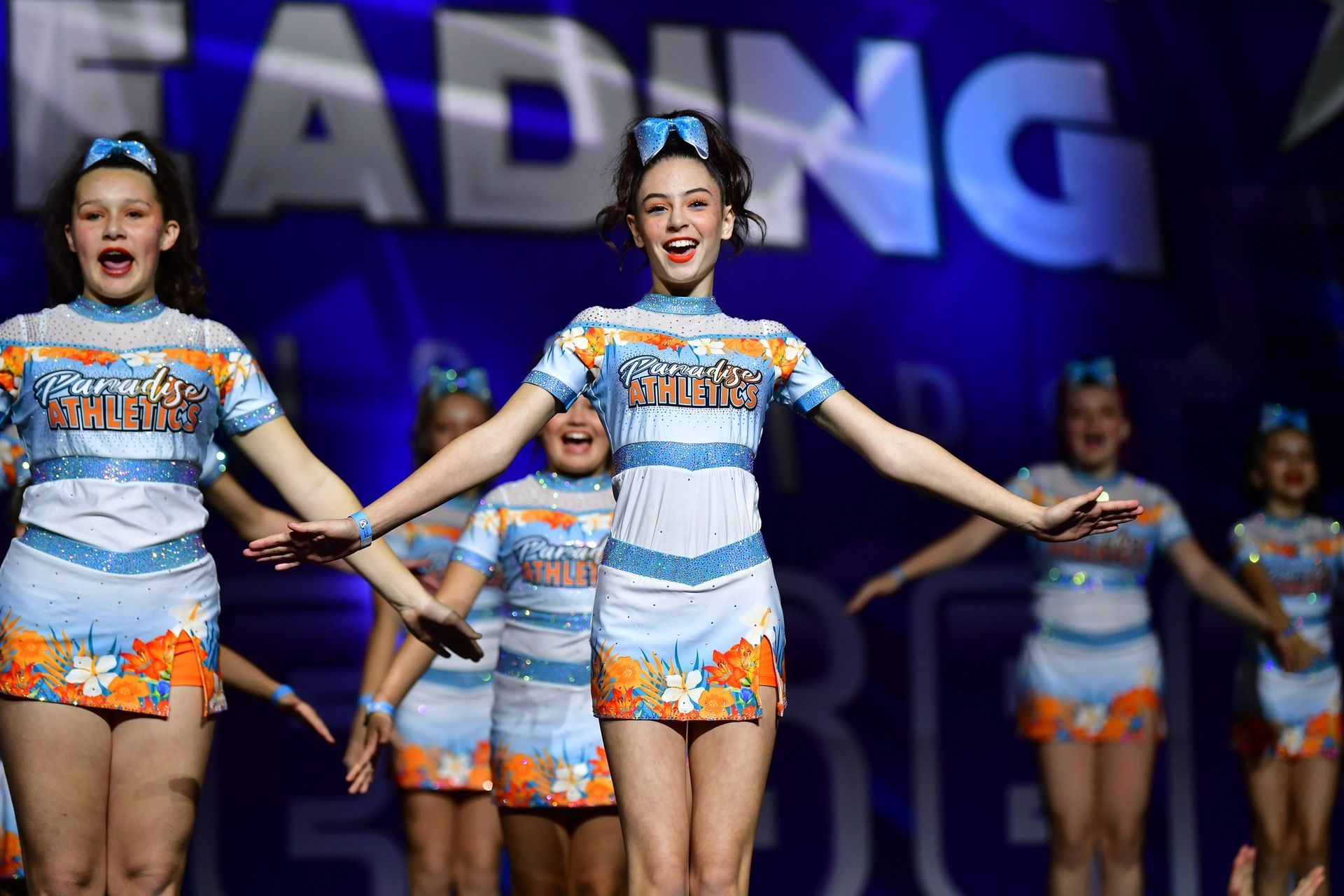 Cheerleaders in light blue and orange floral uniforms performing on a stage with a blue background.