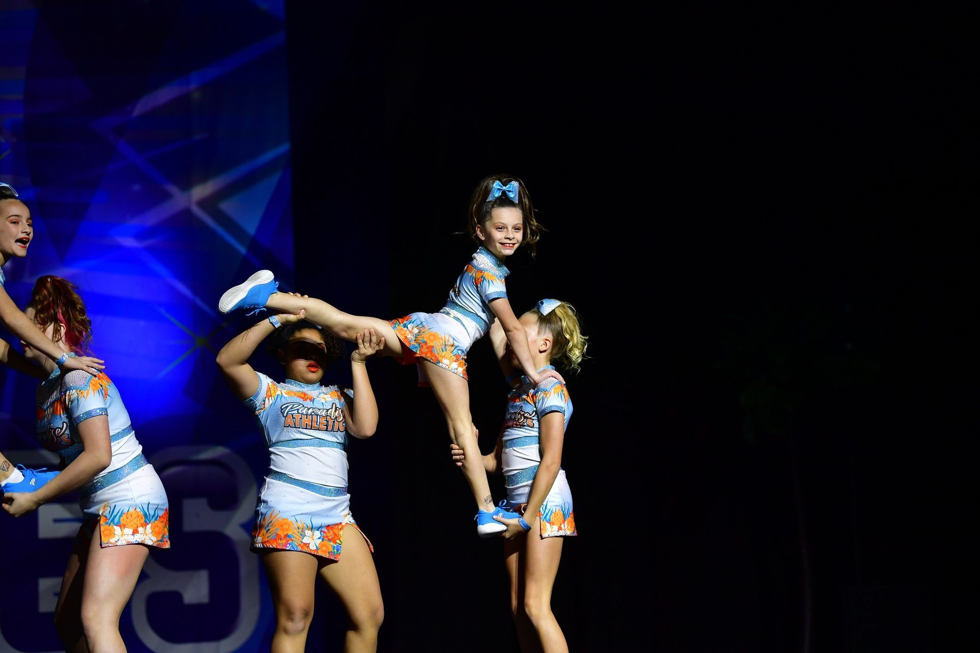 A cheerleading squad in matching light blue and orange uniforms performs a stunt on stage under spotlights.