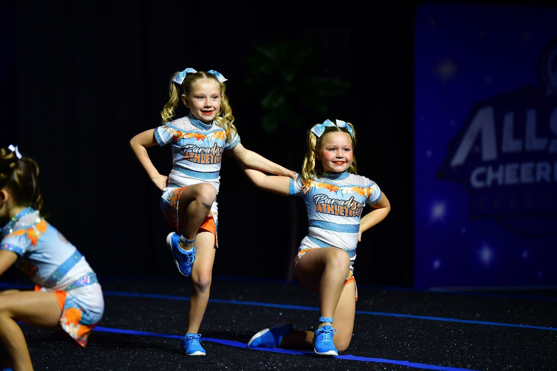 Three cheerleaders in matching blue and orange uniforms perform a routine on a dark stage with a blue event backdrop.