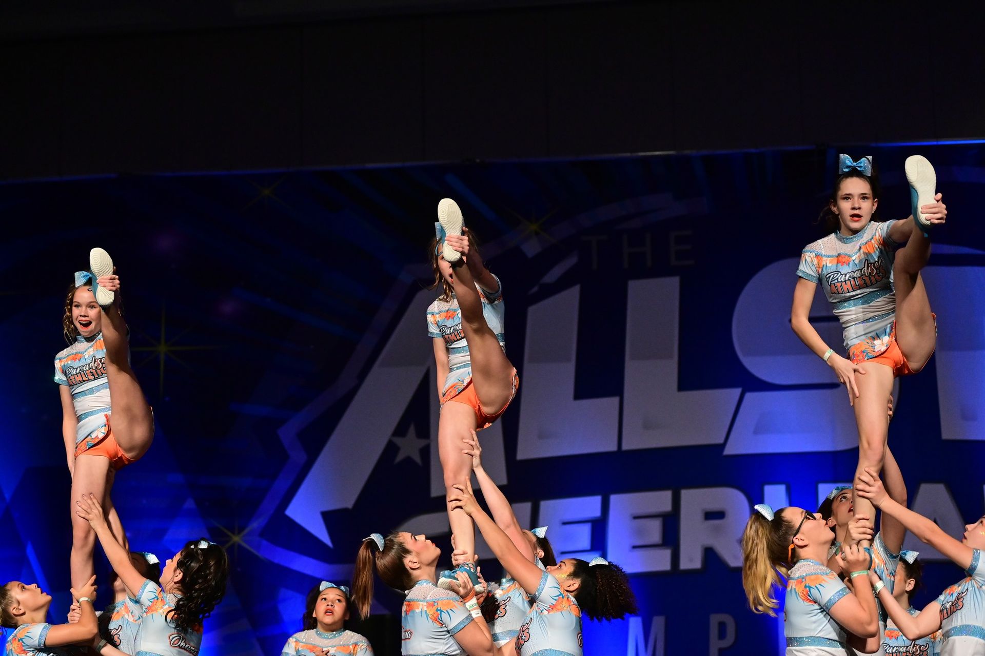 Cheerleaders in matching light blue and orange uniforms perform a coordinated stunt on stage at a competition.